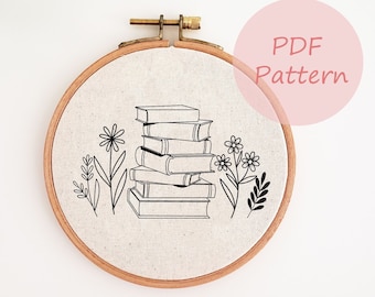 May include: An embroidery hoop with a beige fabric. It features a black line drawing of a stack of books and floral elements. The text "PDF Pattern" is in a pink circle. The hoop is made of wood.