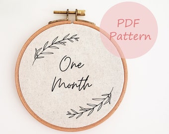 May include: A round embroidery hoop with a beige fabric center. The text "One Month" is written in black script, with delicate leaf sprigs. The hoop is made of wood and has a brass clasp. A pink circle with the text "PDF Pattern" is in the background.