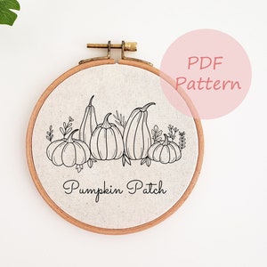 Puede incluir: Un dibujo en blanco y negro de calabazas y hojas sobre un fondo de tela beige. La tela está estirada sobre un aro de bordado de madera. El texto "Pumpkin Patch" está escrito debajo de las calabazas.