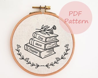 May include: A black and white embroidery pattern of a stack of books with the words "One More Chapter" on the covers. The books are decorated with a small floral design. The pattern is displayed on a round piece of fabric in a wooden embroidery hoop.