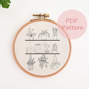 Pode incluir: Um padrão de bordado em preto e branco de uma prateleira com plantas em vasos e livros. O padrão está sobre um fundo de tecido bege e está dentro de um aro de bordado de madeira redondo. O texto "PDF Pattern" está em um círculo rosa no canto superior direito da imagem.