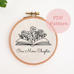 Peut inclure: Un cerceau de broderie blanc avec un dessin au trait noir d'un livre ouvert avec des fleurs qui en sortent. Le texte "One More Chapter" est écrit sous le livre.