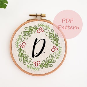 May include: A round embroidery hoop with a cream-colored fabric featuring a green and red wreath design. The wreath surrounds a large black letter "D". The text "PDF Pattern" is in a pink circle in the upper right corner.