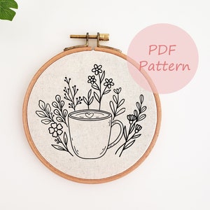 May include: A black and white embroidery pattern of a coffee cup with a heart in the foam and flowers around it. The pattern is in a wooden embroidery hoop.