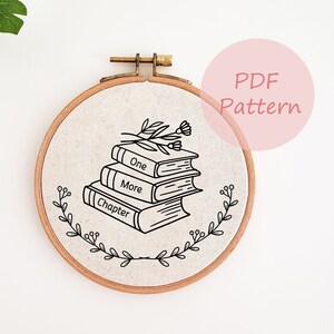 Peut inclure: Un motif de broderie noir et blanc représentant une pile de livres avec les mots "One More Chapter" sur les couvertures. Les livres sont décorés d'un petit motif floral. Le motif est affiché sur un morceau de tissu rond dans un cerceau de broderie en bois.