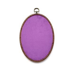 May include: Oval embroidery hoop with a vibrant purple fabric center. The wooden frame has a decorative pattern and a metal loop at the top for hanging. The hoop is set against a white background.