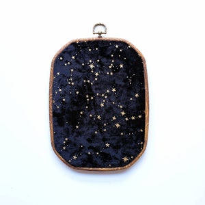 May include: A rectangular wall hanging featuring a dark blue velvet panel decorated with gold stars. The panel is set within a wooden frame and has a brass-coloured metal loop at the top. The stars are arranged in constellations.