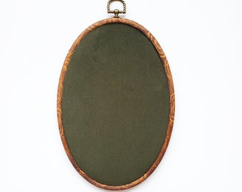 May include: Oval embroidery hoop with a dark green fabric center. The wooden frame has a decorative grain pattern and a brass-colored hanging loop at the top. The hoop is set against a white background.
