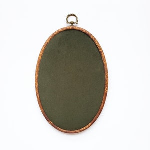 May include: Oval embroidery hoop with a dark green fabric center. The wooden frame has a decorative grain pattern and a brass-colored hanging loop at the top. The hoop is set against a white background.
