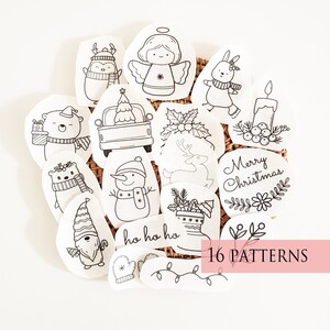 May include: A collection of 16 Christmas-themed patterns, each featuring a unique black and white illustration. Designs include a penguin, angel, bunny, bear, snowman, and festive text. The patterns are arranged on a woven surface.
