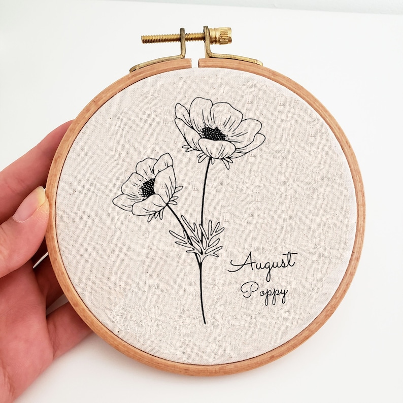 May include: A white embroidery hoop with a black line drawing of two poppy flowers and the text "August Poppy".