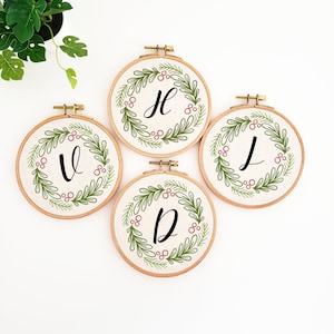 May include: Four embroidery hoops with a cream-colored fabric background. Each hoop features a green wreath design with red berries and a single black letter: V, H, J, and D. The hoops are displayed on a white surface.