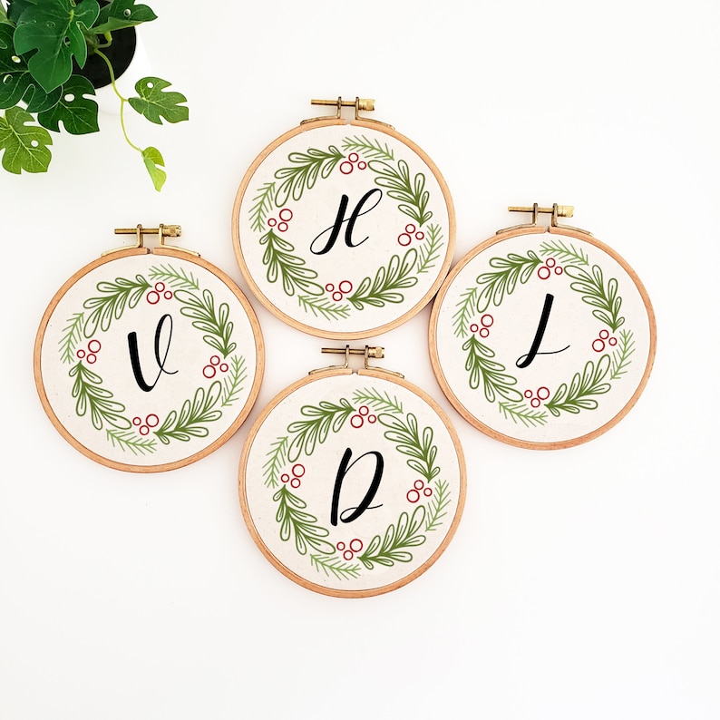 May include: Four embroidery hoops with a cream-colored fabric background. Each hoop features a green wreath design with red berries and a single black letter: V, H, J, and D. The hoops are displayed on a white surface.