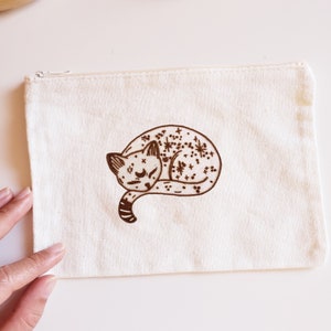 May include: A cream-colored canvas pouch with a zipper closure. The pouch features a brown illustration of a sleeping cat with star and dot patterns. The cat is curled up, and the design is centered on the pouch.