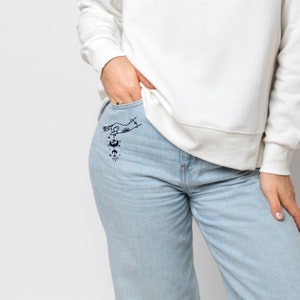 May include: Light blue jeans with a black line art design on the pocket. The design features a hand, stars, and celestial elements. The person is wearing a white sweatshirt.