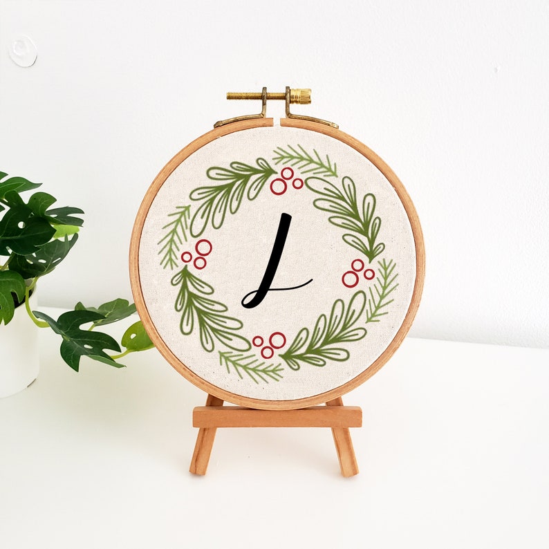 May include: A round embroidery hoop featuring a wreath design with green leaves and red berries. The letter "L" is in the center. The hoop is displayed on a small wooden easel, perfect for home decor.