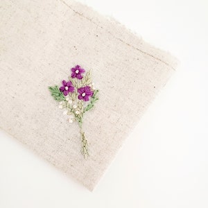 May include: A beige linen fabric with a hand-embroidered floral design. The embroidery features purple flowers, green leaves, and small white bead-like accents. The fabric has a raw edge on the top right corner.