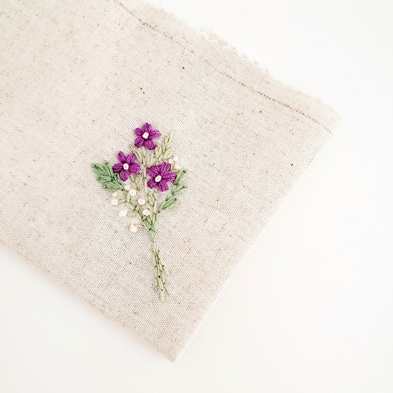 May include: A beige linen fabric with a hand-embroidered floral design. The embroidery features purple flowers, green leaves, and small white bead-like accents. The fabric has a raw edge on the top right corner.