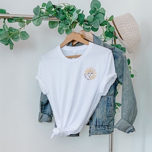 May include: White t-shirt with a gold embroidered sun and moon design. The t-shirt is hanging on a wooden hanger, with a denim jacket and a straw hat in the background. The shirt is tied at the waist.