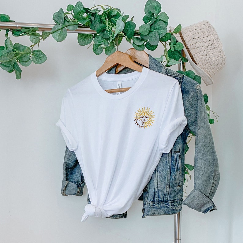 May include: White t-shirt with a gold embroidered sun and moon design. The t-shirt is hanging on a wooden hanger, with a denim jacket and a straw hat in the background. The shirt is tied at the waist.