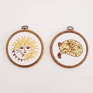 May include: Two round embroidery hoops with wooden frames. One features a yellow sun with a face and the moon. The other depicts a sleeping cat with star details. Both are on white fabric.