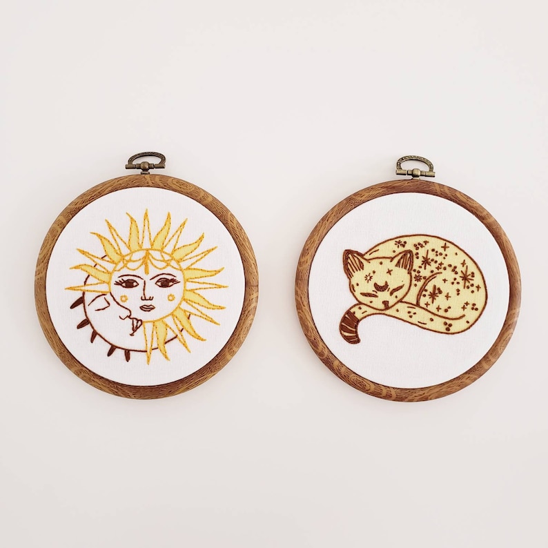 May include: Two round embroidery hoops with wooden frames. One features a yellow sun with a face and the moon. The other depicts a sleeping cat with star details. Both are on white fabric.
