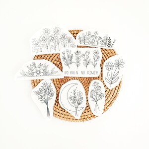 May include: A collection of black and white floral stickers arranged on a woven wicker mat. The stickers feature various flower designs, including daisies and bouquets, with the phrase "NO RAIN NO FLOWER" on one. The stickers are in a variety of shapes.