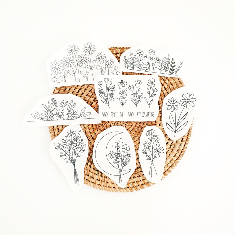 May include: A collection of black and white floral stickers arranged on a woven wicker mat. The stickers feature various flower designs, including daisies and bouquets, with the phrase "NO RAIN NO FLOWER" on one. The stickers are in a variety of shapes.