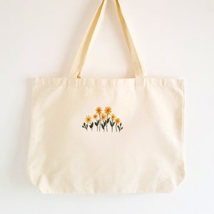 May include: Cream-colored tote bag with embroidered yellow and orange flowers. The bag has two long handles and is made of a sturdy fabric. The floral design is centered on the front of the bag.