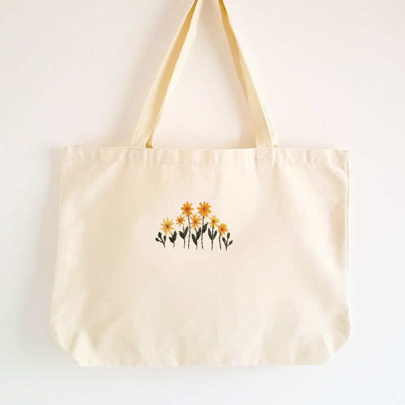 May include: Cream-colored tote bag with embroidered yellow and orange flowers. The bag has two long handles and is made of a sturdy fabric. The floral design is centered on the front of the bag.