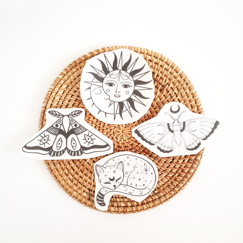 May include: Four celestial-themed fabric patches: a sun and moon face, two moths, and a sleeping cat with stars. The patches are black and white, placed on a round, woven, light brown placemat. The background is white.
