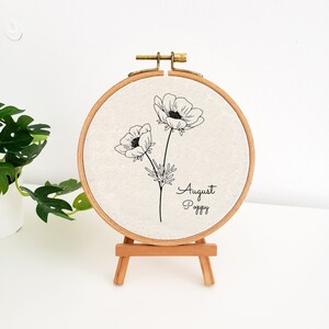 May include: A white embroidery hoop with a black line drawing of two poppy flowers and the text "August Poppy". The hoop is on a small wooden easel.