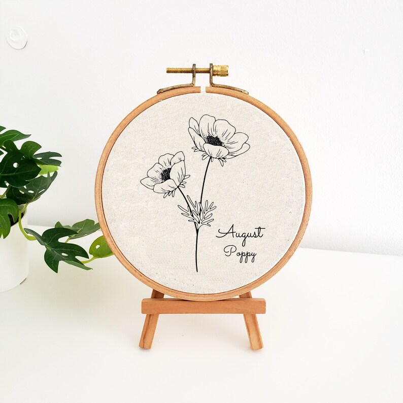 May include: A white embroidery hoop with a black line drawing of two poppy flowers and the text "August Poppy". The hoop is on a small wooden easel.