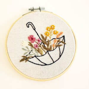 May include: Embroidered artwork in a wooden hoop. The design features a black outline of an umbrella filled with colorful flowers in shades of pink, yellow, and orange, with green and brown foliage. The background is a light beige fabric.