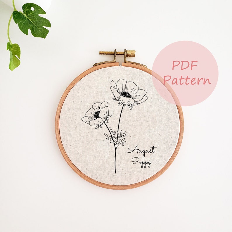 May include: A black and white line drawing of an August Poppy flower on a white fabric background in a wooden embroidery hoop. The text "August Poppy" is written below the flower.