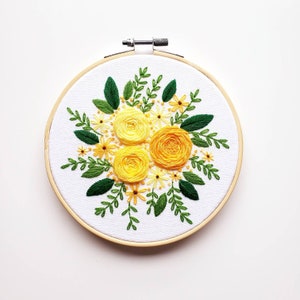 May include: Hand-stitched embroidery art featuring a vibrant floral design. The artwork showcases three large yellow roses surrounded by smaller yellow flowers and green leaves. The embroidery is set in a 6-inch wooden hoop, perfect for wall decor.