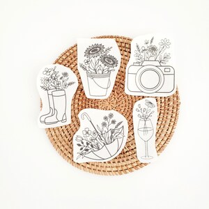 May include: A collection of six white, die-cut shapes featuring black line drawings of flowers in various containers. The shapes include a bucket, camera, wine glass, umbrella, and rain boots, all arranged on a woven, circular mat.