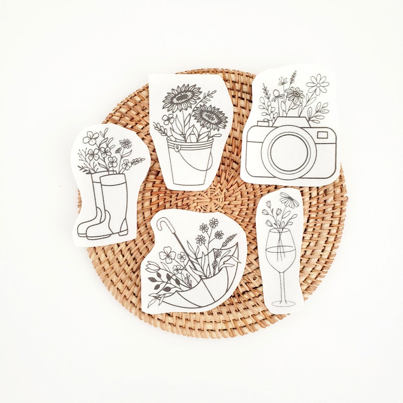 May include: A collection of six white, die-cut shapes featuring black line drawings of flowers in various containers. The shapes include a bucket, camera, wine glass, umbrella, and rain boots, all arranged on a woven, circular mat.