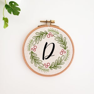 May include: A round embroidery hoop with a beige fabric featuring a green and red wreath design. The wreath surrounds the black letter "D". The wooden hoop is held together with a brass clasp. A decorative piece for the home.