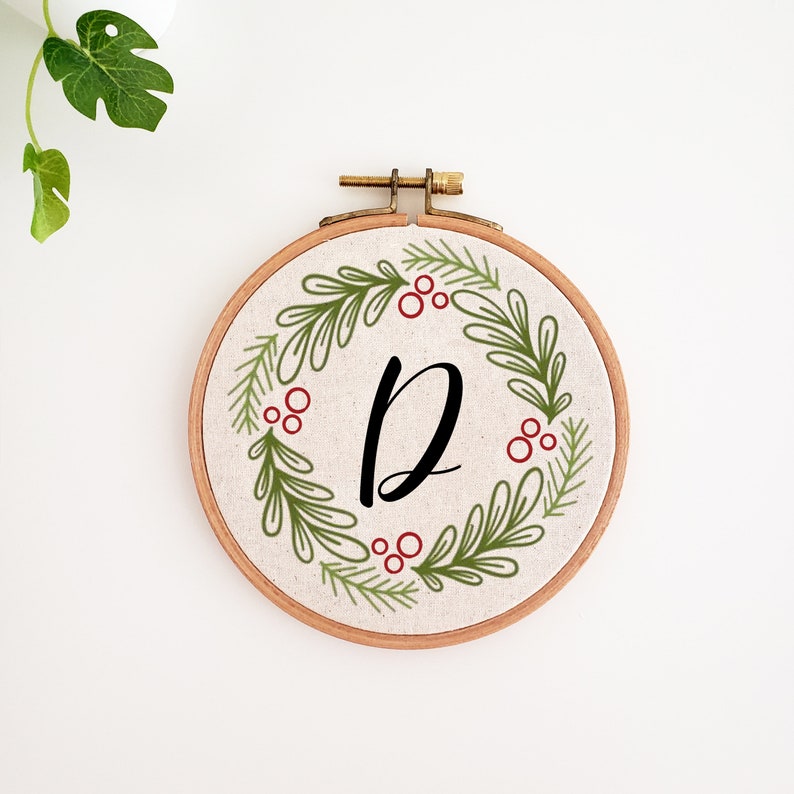 May include: A round embroidery hoop with a beige fabric featuring a green and red wreath design. The wreath surrounds the black letter "D". The wooden hoop is held together with a brass clasp. A decorative piece for the home.