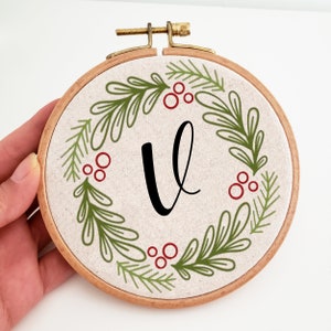 May include: A round embroidery hoop with a beige fabric featuring a green and red wreath design. The wreath surrounds a large black letter "V". The wooden hoop has a brass clasp.