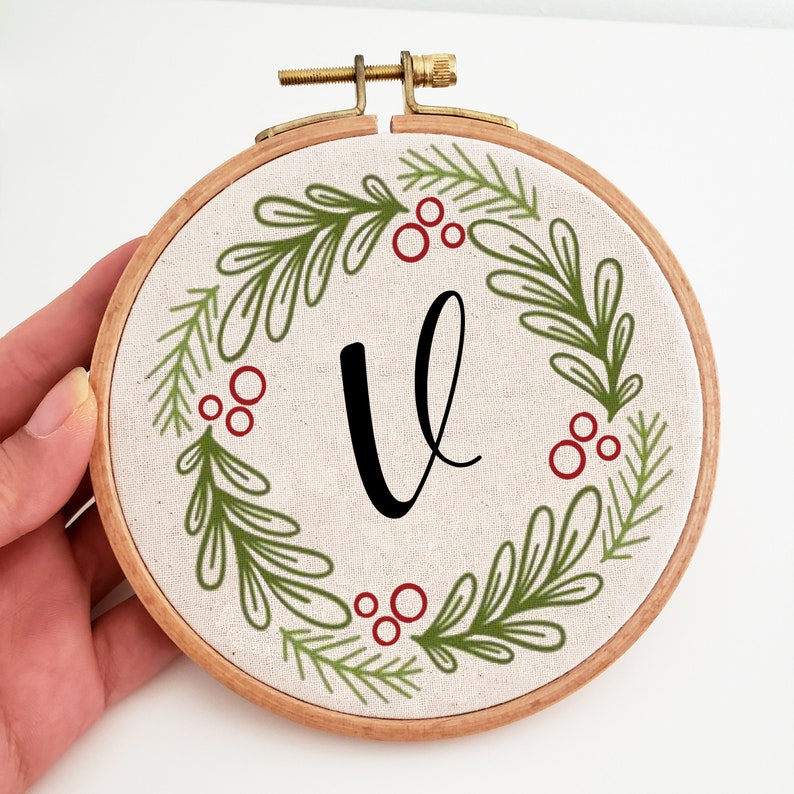 May include: A round embroidery hoop with a beige fabric featuring a green and red wreath design. The wreath surrounds a large black letter "V". The wooden hoop has a brass clasp.