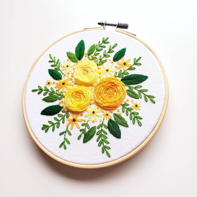 May include: Hand-stitched embroidery art featuring a floral design. The artwork showcases three large yellow roses surrounded by smaller yellow flowers and green leaves. The embroidery is set in a wooden hoop, creating a framed piece of textile art.