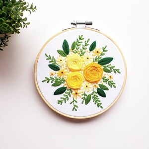May include: Hand-stitched floral embroidery art in a wooden hoop. The design features three large yellow roses surrounded by smaller yellow flowers and green leaves. The embroidery is on a white fabric background.