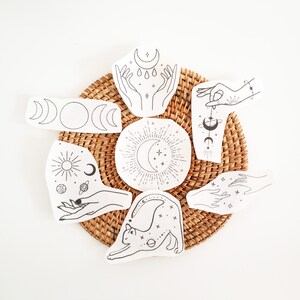 May include: A collection of white fabric patches with black line art illustrations of celestial and mystical themes. Designs include moons, hands, suns, planets, and a stretching cat. The patches are arranged on a woven, circular mat.