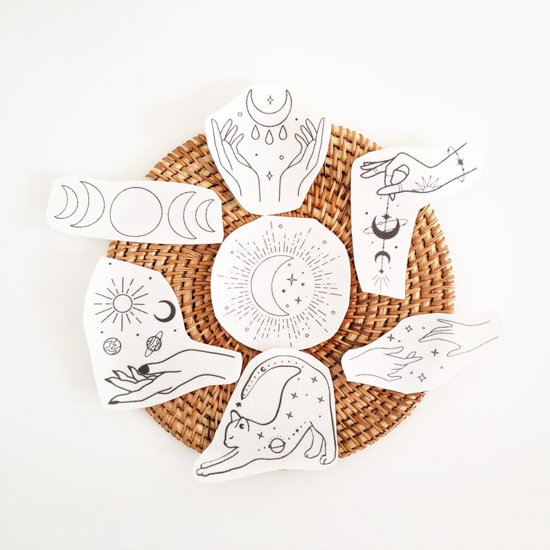 May include: A collection of white fabric patches with black line art illustrations of celestial and mystical themes. Designs include moons, hands, suns, planets, and a stretching cat. The patches are arranged on a woven, circular mat.