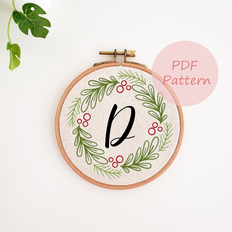 May include: A round embroidery hoop with a cream-colored fabric featuring a green and red wreath design. The wreath surrounds a large black letter "D". The text "PDF Pattern" is in a pink circle in the upper right corner.