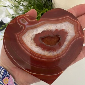 Pu&ograve; includere: Pietra di agata a forma di cuore con finitura lucida e lucente. La pietra presenta bande concentriche di rosso intenso e bianco, con una cavit&agrave; centrale di cristalli scintillanti. La pietra naturale &egrave; tenuta in mano.