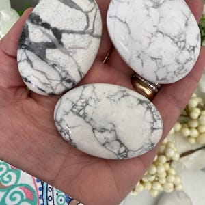May include: Three white Howlite stones with gray veining are held in a hand. The stones are oval-shaped and polished, with a smooth surface. The background includes white flowers and a patterned sleeve.