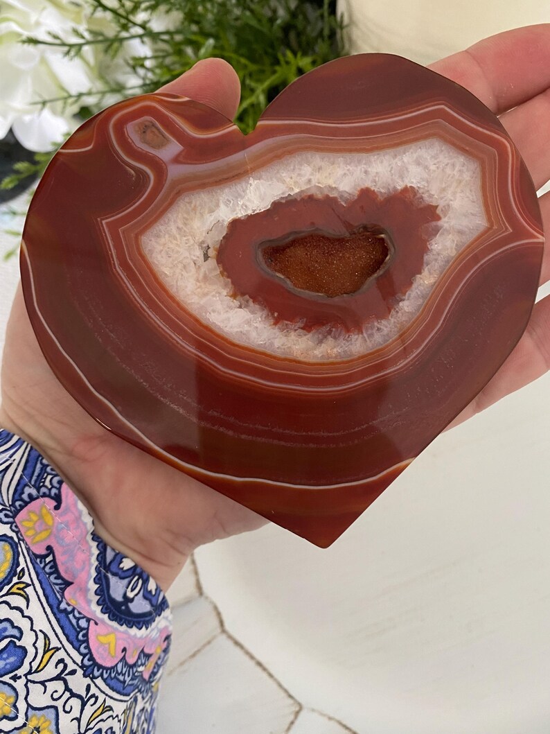 Pu&ograve; includere: Pietra di agata a forma di cuore con finitura lucida e lucente. La pietra presenta bande concentriche di rosso intenso e bianco, con una cavit&agrave; centrale di cristalli scintillanti. La pietra naturale &egrave; tenuta in mano.